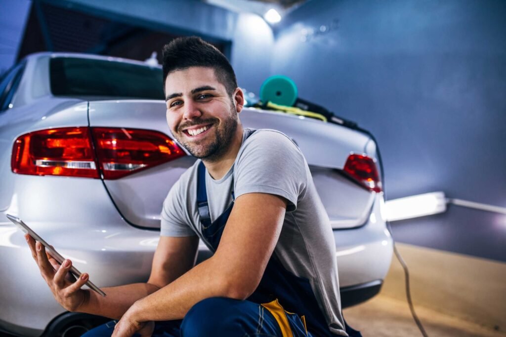 Smiling auto detailer crouches beside the rear of a silver car in a garage, holding a smartphone, with detailing tools resting on the trunk and bright workshop lighting in the background.